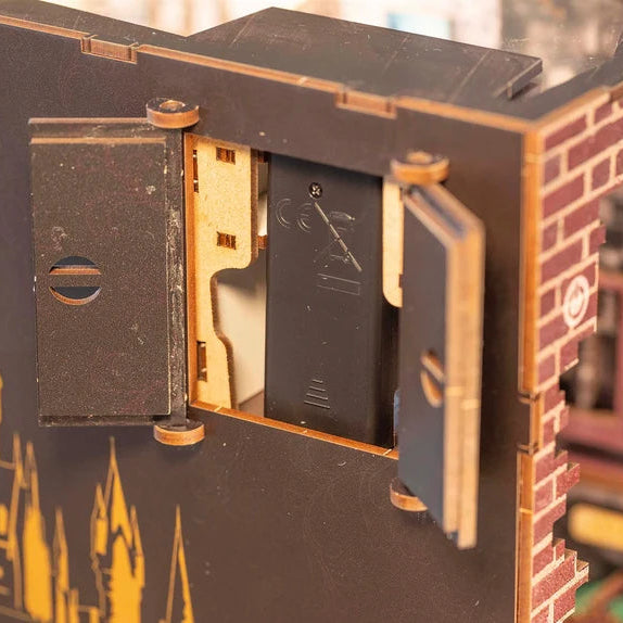 Close-up of a wooden 3D puzzle model of a building, showing the interior battery compartment with a black battery and wooden switches, alongside a brick-patterned wall section.