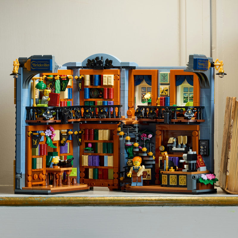 A detailed LEGO model of a two-story library or bookstore, featuring orange bookshelves filled with colorful books, a gray exterior, and a small LEGO minifigure standing in the doorway. The set is photographed on a shelf against a plain indoor background.