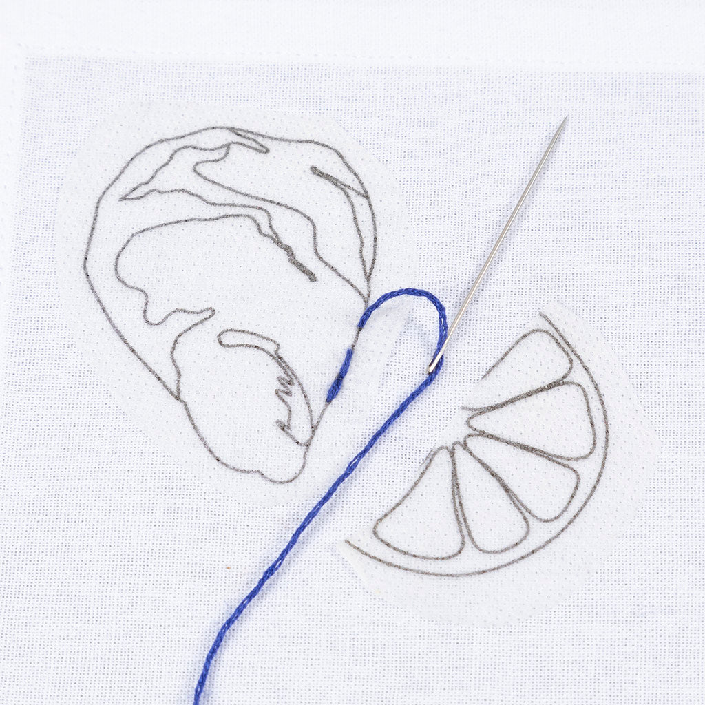 Embroidery design of a hand holding a lemon with a needle and thread on white fabric.