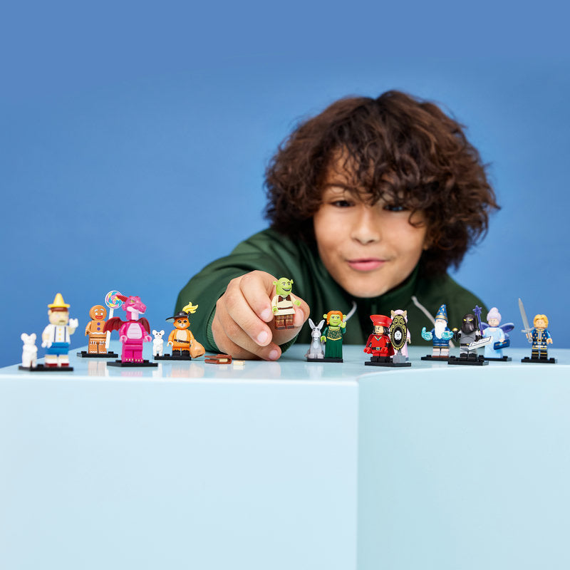 A child with curly brown hair wearing a green shirt smiles while holding a small green character figure, with several other colorful toy minifigures lined up on a light blue surface against a solid blue background.