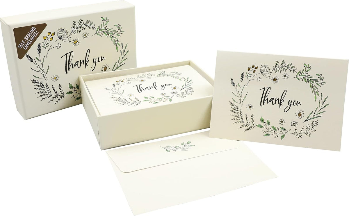 A boxed thank you card set with a floral wreath illustration and the words 'thank you' on the card, shown with an open box, a closed box, and an envelope on a white background.