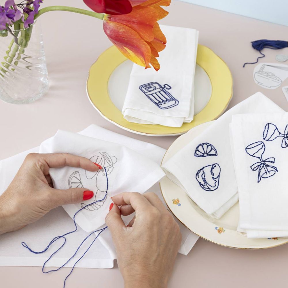 Person stitching on a white fabric with embroidery designs, surrounded by embroidered napkins and flowers.