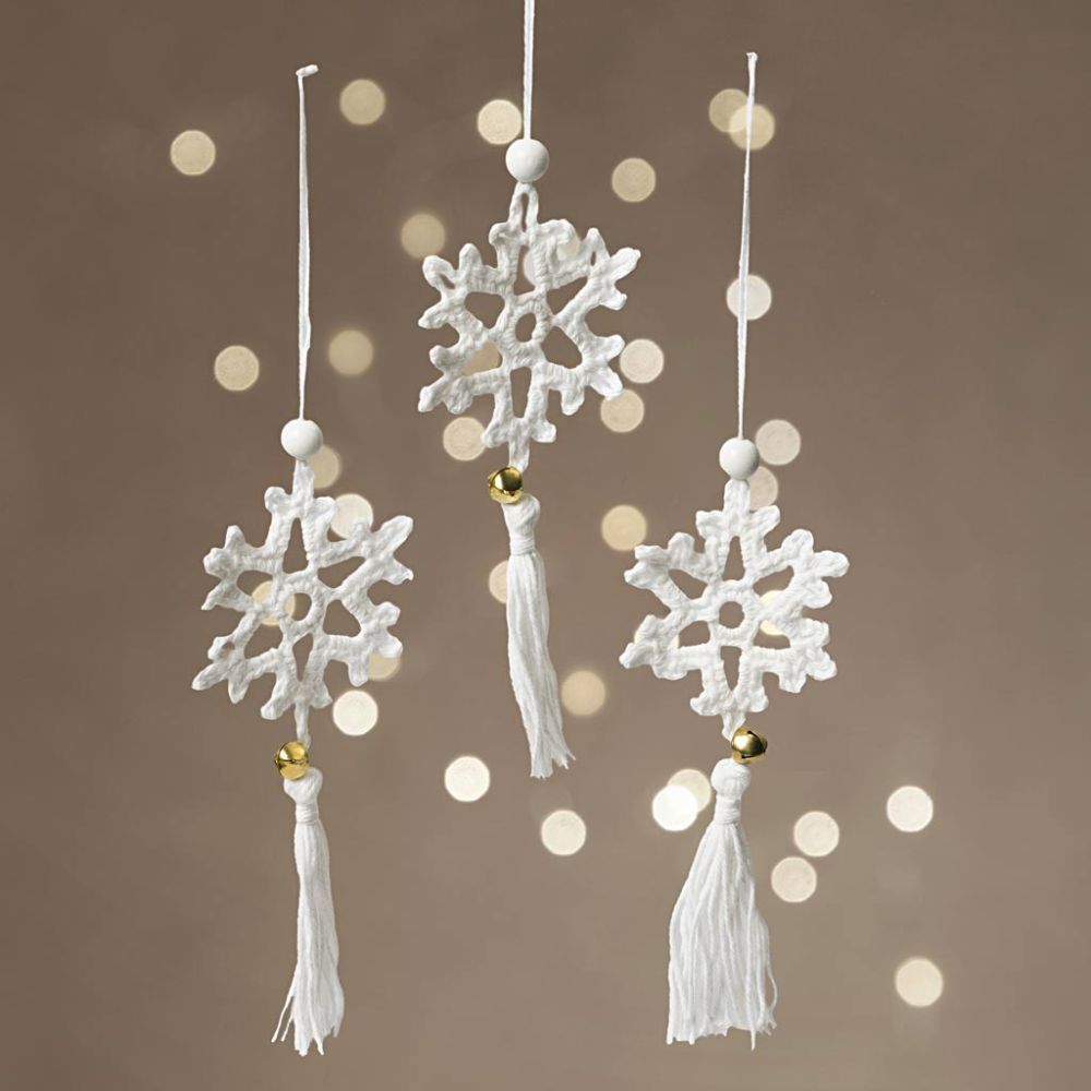 Three white snowflake-shaped ornaments with tassels on a brown background with bokeh lights.
