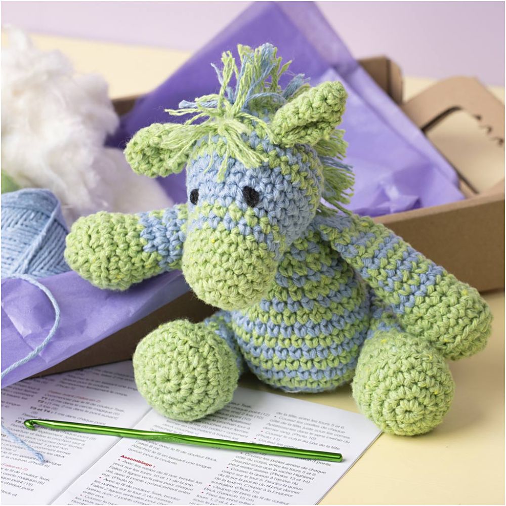 Crocheted green and blue dragon toy with a crochet hook and pattern sheet on a table.