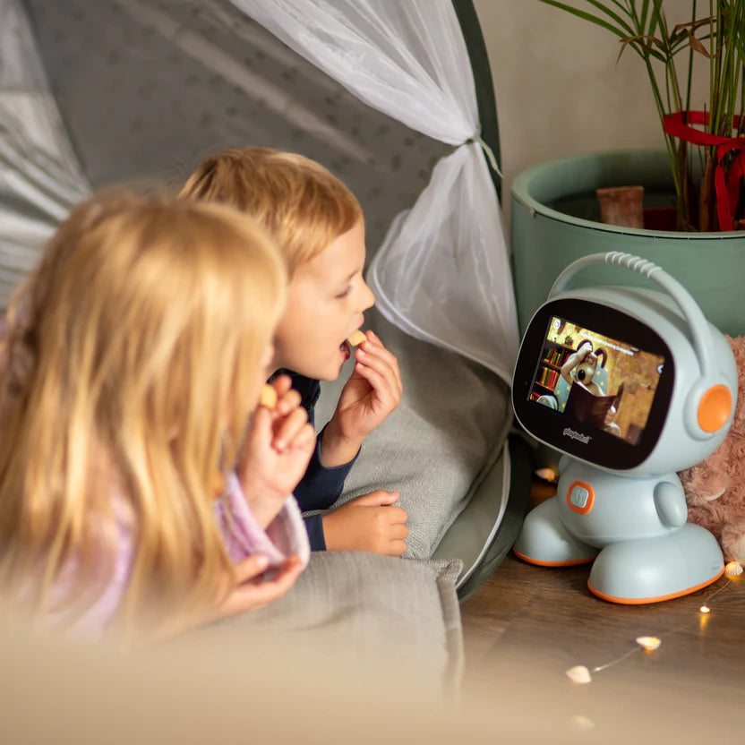 Two children are lying on a floor beside a small robot-like video call toy with a screen, in a cozy indoor setting with a canopy tent and a potted plant in the background.