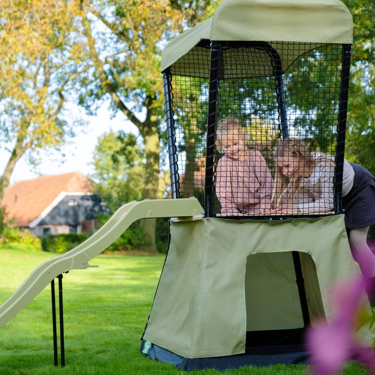Children playing inside a green playhouse with a slide in a park setting.