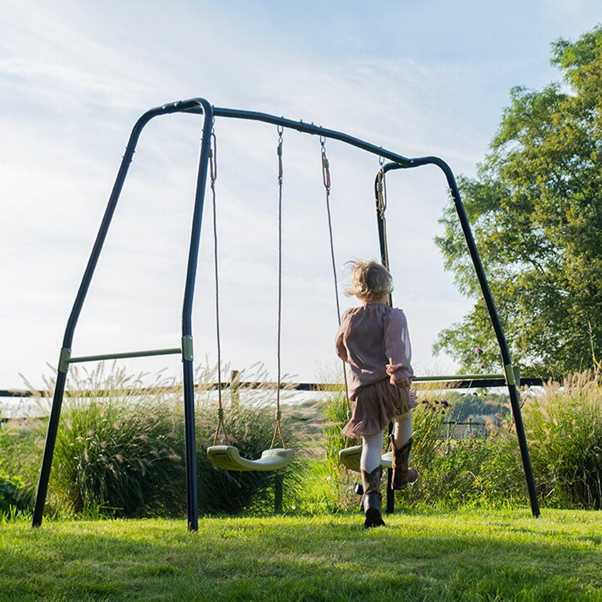 Person standing next to a swing set in a garden with trees and grass in the background