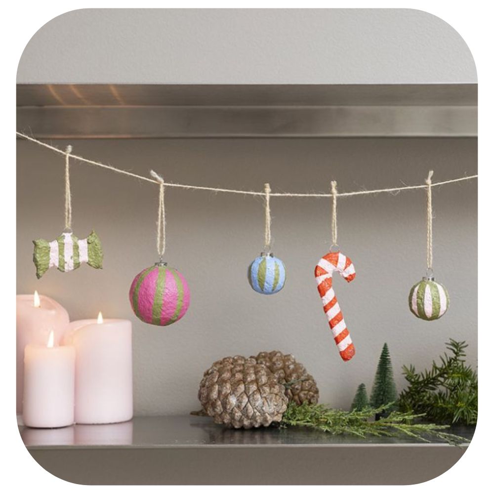 homemade christmas decorations hanging on a string over a shelf with candles, pinecones and green foliage