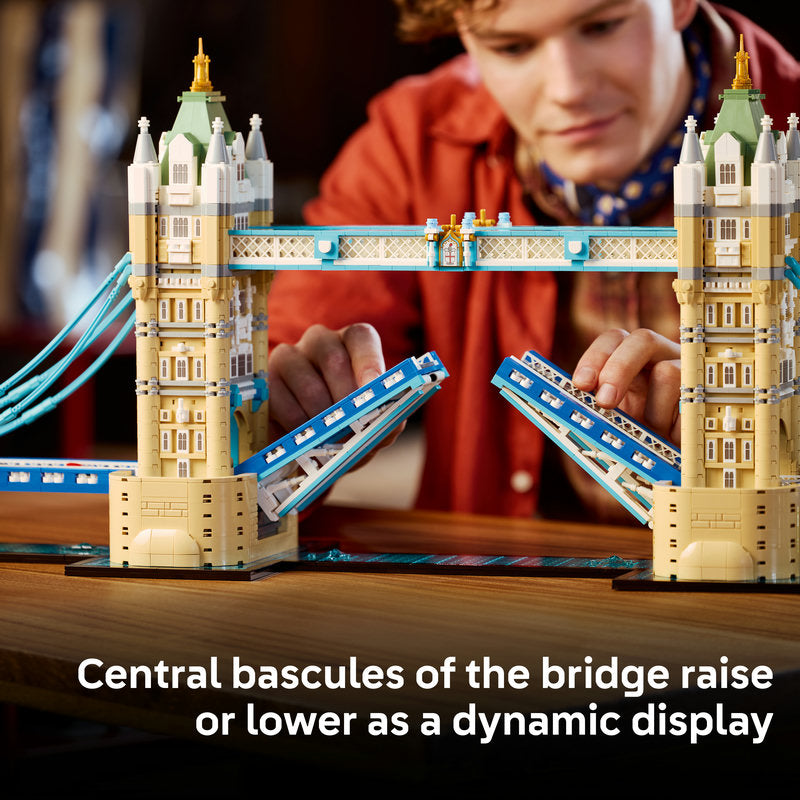 A person in a red jacket is building or playing with a detailed London Tower Bridge model set on a wooden table, with the bridge's central bascules raised. White text at the bottom reads, 'Central bascules of the bridge raise or lower as a dynamic display.
