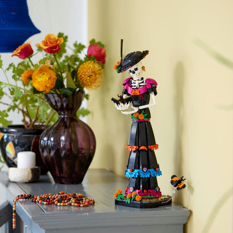 A decorative skeleton figurine dressed in a black dress with colorful floral trim stands on a tabletop beside a dark vase of flowers and a beaded necklace, against a pale yellow wall.