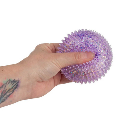 A hand holding a purple textured stress ball filled with small iridescent beads against a plain white background.