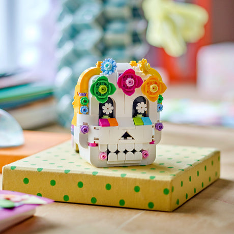 A LEGO Day of the Dead sugar skull building set displayed on top of a yellow gift box with green polka dots, set against a warm, blurred background.