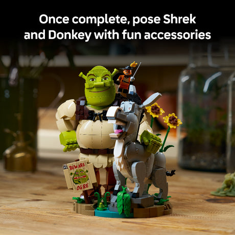 A LEGO-style Shrek and Donkey set displayed on a wooden surface, with the green ogre Shrek and a sign reading 'Beware of Ogre,' and a gray donkey with flowers on its back. White text at the top says, 'Once complete, pose Shrek and Donkey with fun accessories.