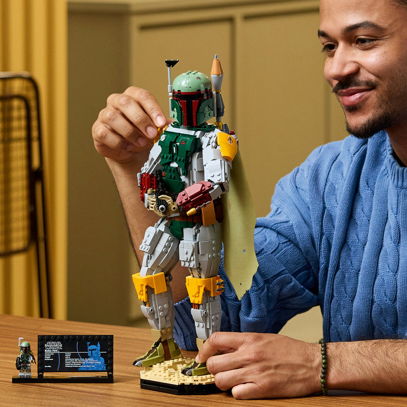 A man in a blue cable-knit sweater holds a detailed LEGO Boba Fett action figure on a tabletop, with a small display card and mini-droid visible nearby in an indoor room setting.