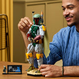 A man in a blue cable-knit sweater holds a detailed LEGO Boba Fett action figure on a tabletop, with a small display card and mini-droid visible nearby in an indoor room setting.