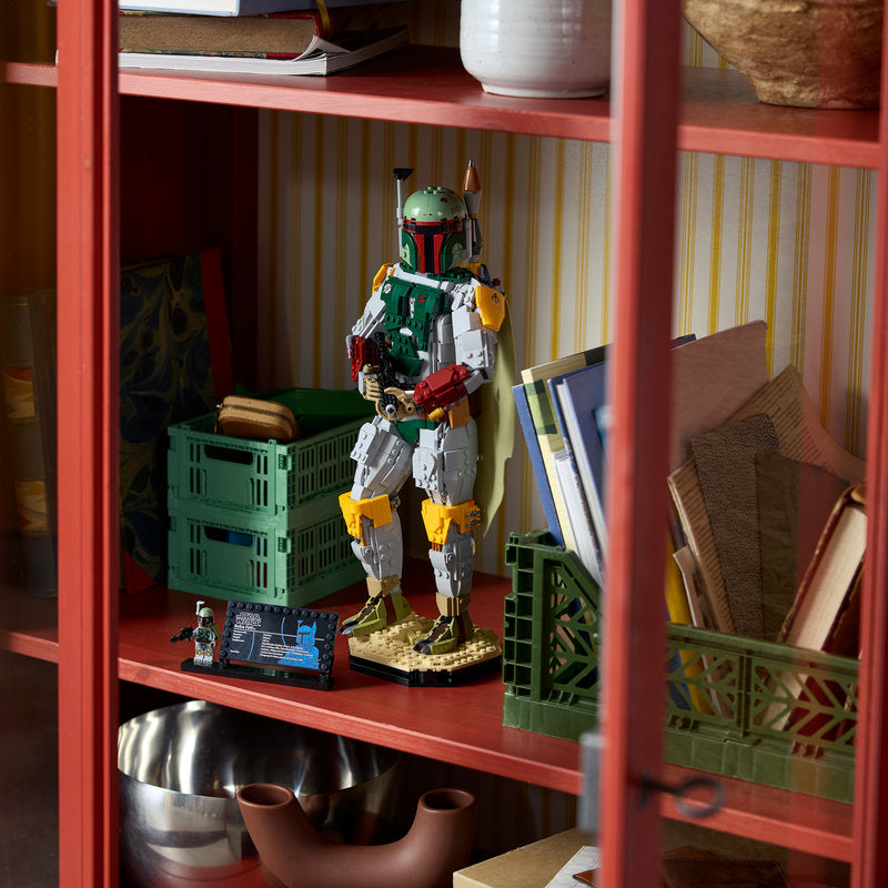 A LEGO Boba Fett toy figure stands on a shelf inside a red wooden cabinet, surrounded by books, green storage crates, and household decor.