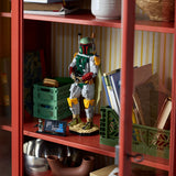 A LEGO Boba Fett toy figure stands on a shelf inside a red wooden cabinet, surrounded by books, green storage crates, and household decor.