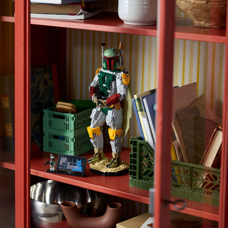 A LEGO Boba Fett toy figure stands on a shelf inside a red wooden cabinet, surrounded by books, green storage crates, and household decor.