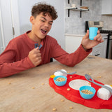 A young man with curly brown hair wearing a rust-red long-sleeve shirt sits at a wooden table in a kitchen setting, holding up a small light blue cup. In front of him is a red play food tray with toy cereal bowls, spoons, and plates.