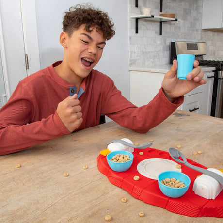 A young man with curly brown hair wearing a rust-red long-sleeve shirt sits at a wooden table in a kitchen setting, holding up a small light blue cup. In front of him is a red play food tray with toy cereal bowls, spoons, and plates.