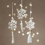 Three white snowflake-shaped ornaments with tassels on a brown background with bokeh lights.