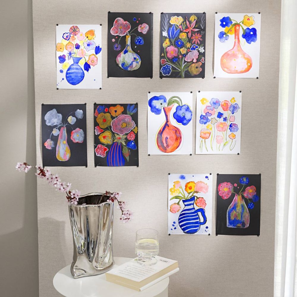 Colourful flowers in a silver vase in front of paintings on a wall above a table.