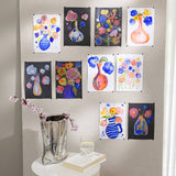 Colourful flowers in a silver vase in front of paintings on a wall above a table.