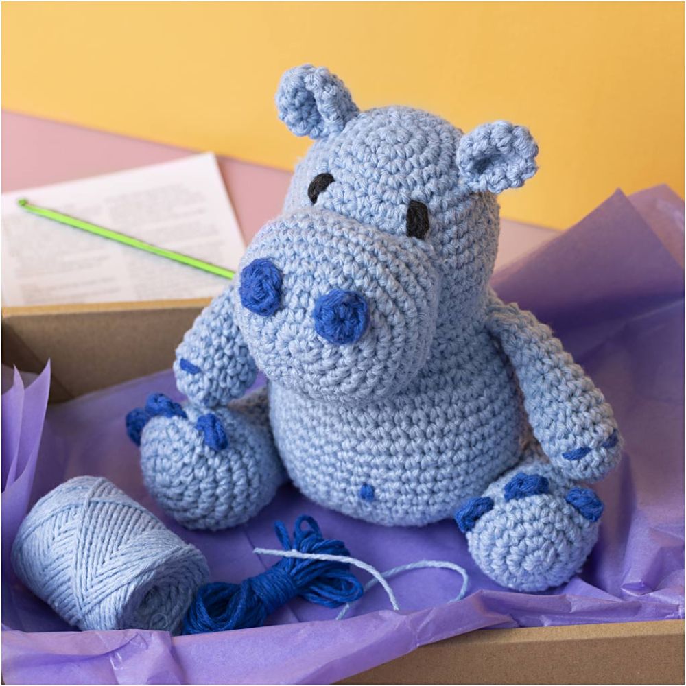 Crocheted hippo toy with yarn and a needle on purple tissue paper against a yellow background