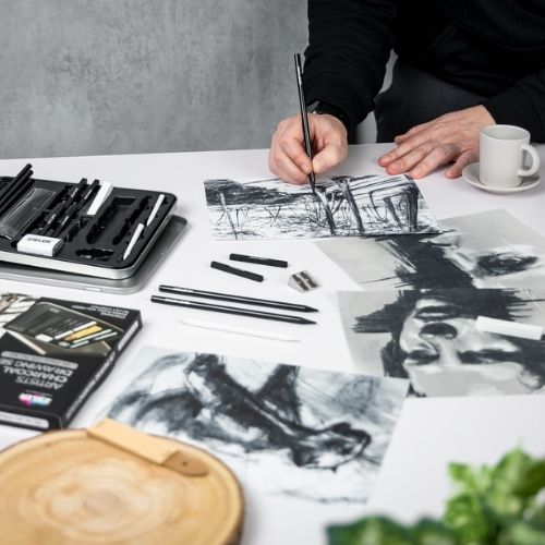 an artist hands drawing with charcoal pencils next to other drawings on a table