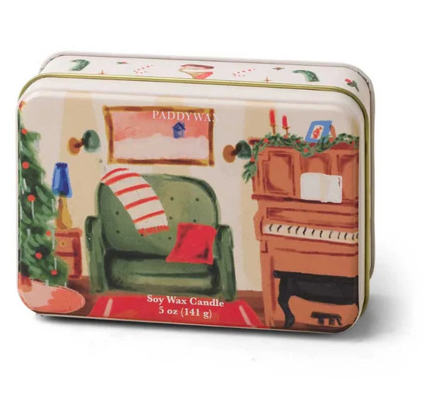 A rectangular tin candle with a festive illustrated design showing a cozy living room scene with a green armchair, red striped throw, piano, and Christmas tree. The front text reads 'PADDYWAX' and 'Soy Wax Candle 5 oz (141 g)'.