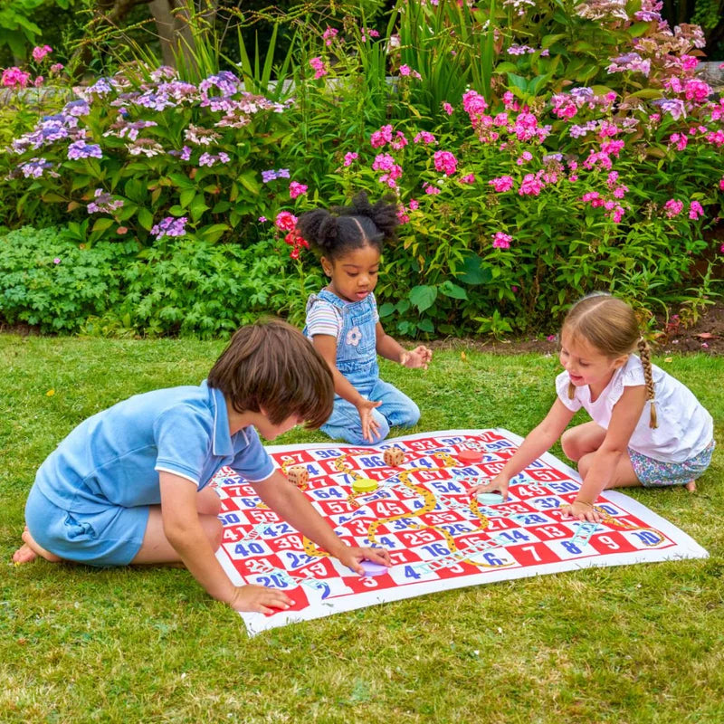 Three children playing with a large snakes and ladders game on grass with flowers in the background