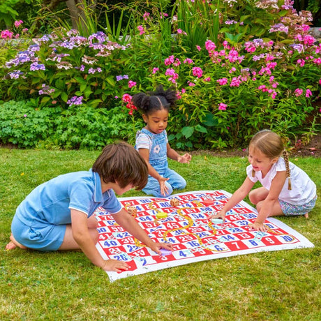 Three children playing with a large snakes and ladders game on grass with flowers in the background