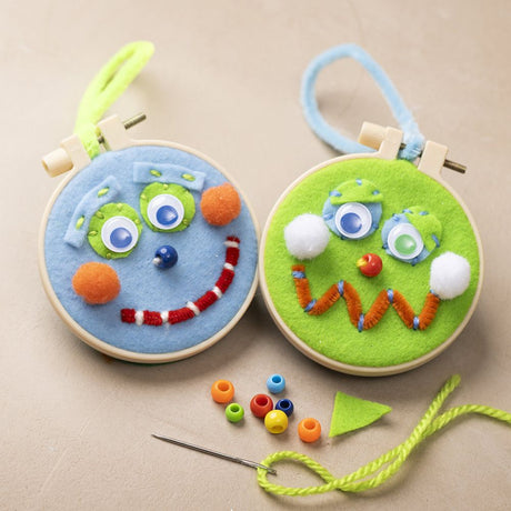 Two felt and embroidered face designs in wooden hoops with colorful yarn on a beige background.