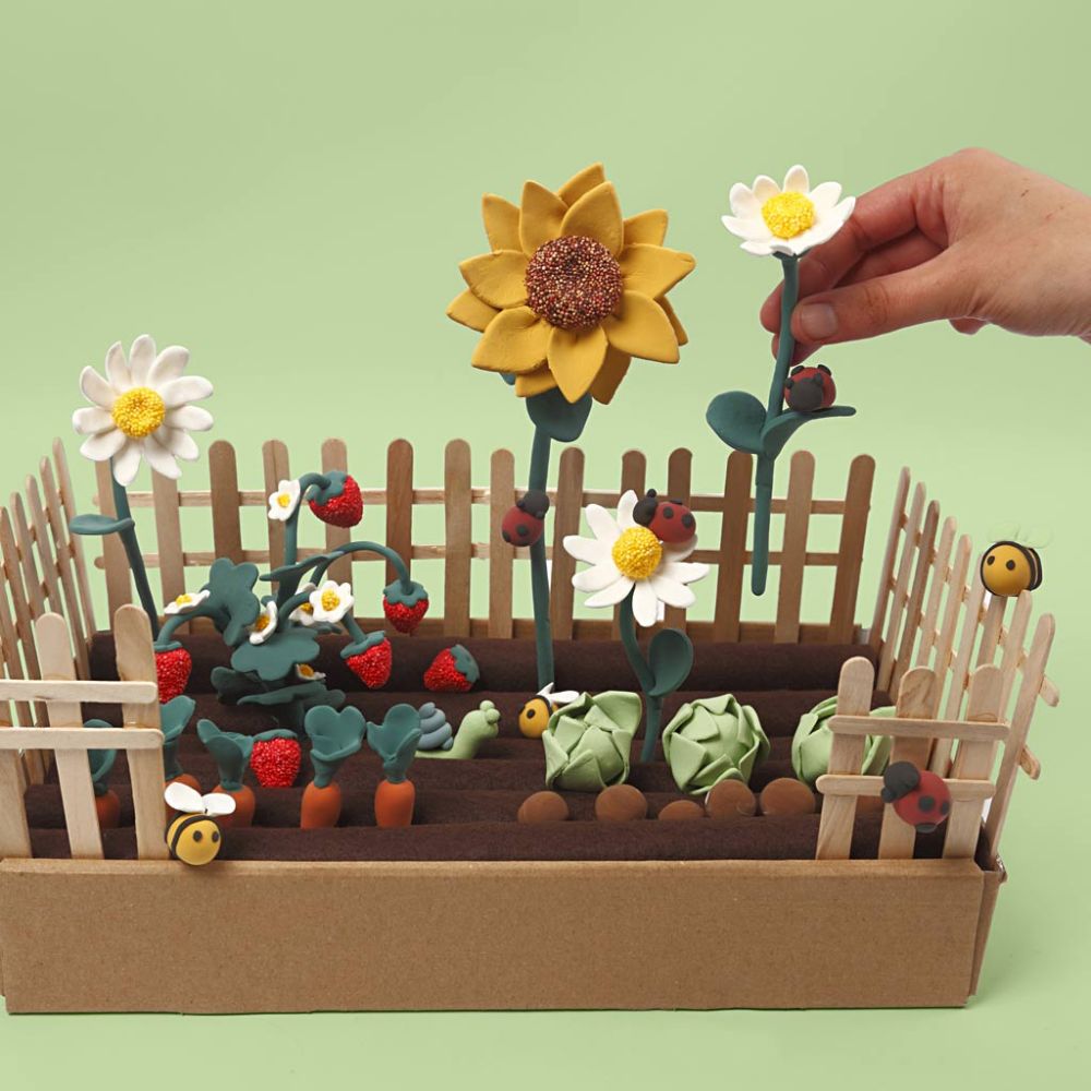 A garden plot made from popsicles and cardboard filled with yellow and white flowers, strawberries and bumblebees made with clay