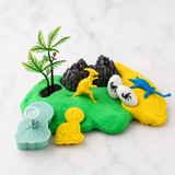 A dinosaur-themed play dough cookie cutter set on a white marble background, featuring a mint green dinosaur-shaped cutter, yellow and blue dinosaur figures, a green fern, black rocks, and white eggs.