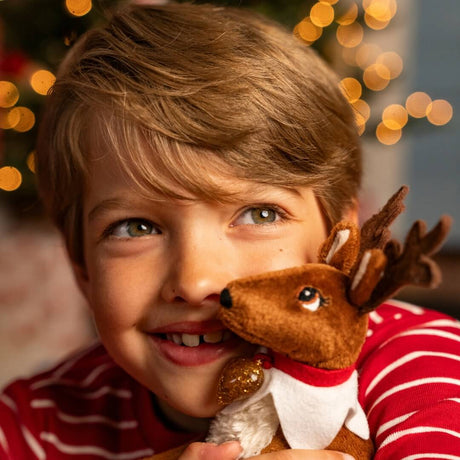A young boy with light brown hair smiles while holding a brown plush reindeer toy with antlers and a red-and-white outfit, set against a softly blurred Christmas tree with warm lights.