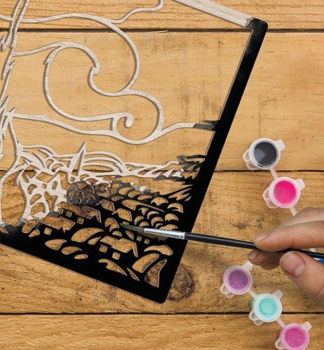 Hand painting a black cutout design on a wooden surface with small containers of paint.