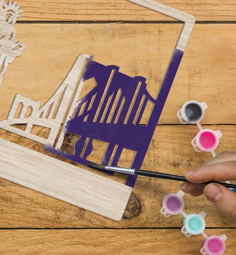 Person painting a wooden cutout of a chair with purple paint on a wooden surface.