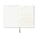 An open bullet journal with a light blue cover and cream-colored interior pages, showing grid sections on the left and calendar/plan inserts on the right, with a gold ribbon bookmark hanging from the bottom.