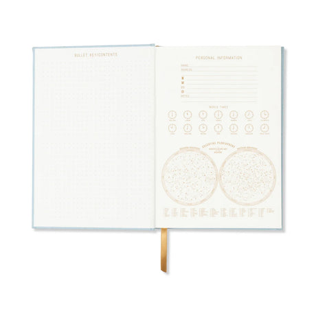 An open bullet journal with a light blue cover and cream-colored interior pages, showing grid sections on the left and calendar/plan inserts on the right, with a gold ribbon bookmark hanging from the bottom.