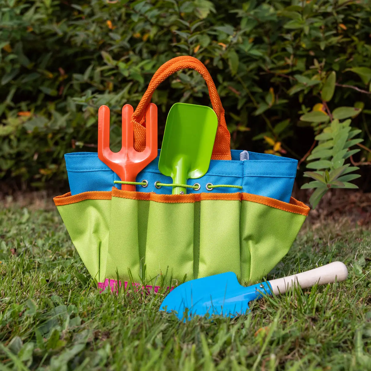 Colorful gardening tools in a green and blue bag on grass with plants in the background