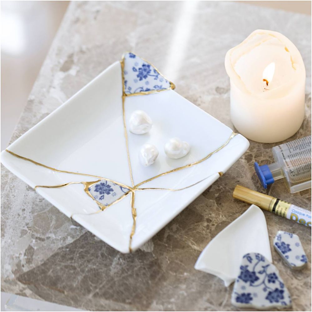White porcelain dish with gold accents and blue floral patterns, accompanied by a candle and small containers on a marble surface.