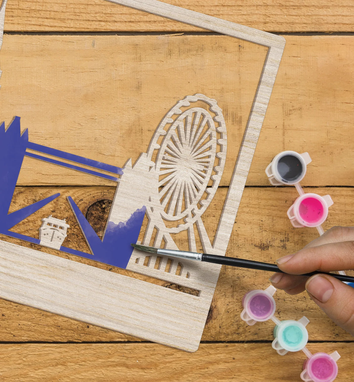 Person painting a wooden cutout of a Ferris wheel with a brush on a wooden surface.