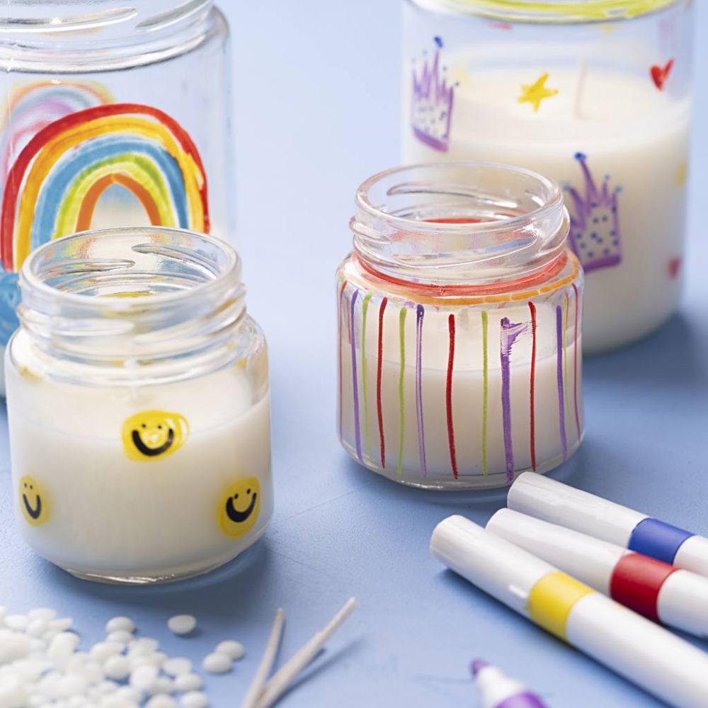 homemade candles in glass jars painted with smiley faces and rainbows on a blue table with craft supplies