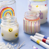 homemade candles in glass jars painted with smiley faces and rainbows on a blue table with craft supplies