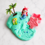 A small mermaid-themed clay or play-dough island on a white marble surface, featuring turquoise base color with a red-haired mermaid figure, a yellow fish character, palm trees, a pink starfish-shaped mold, and small multicolored embellishments.