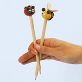 Hand holding two wooden pencils with ladybug and bee toppers on a light blue background