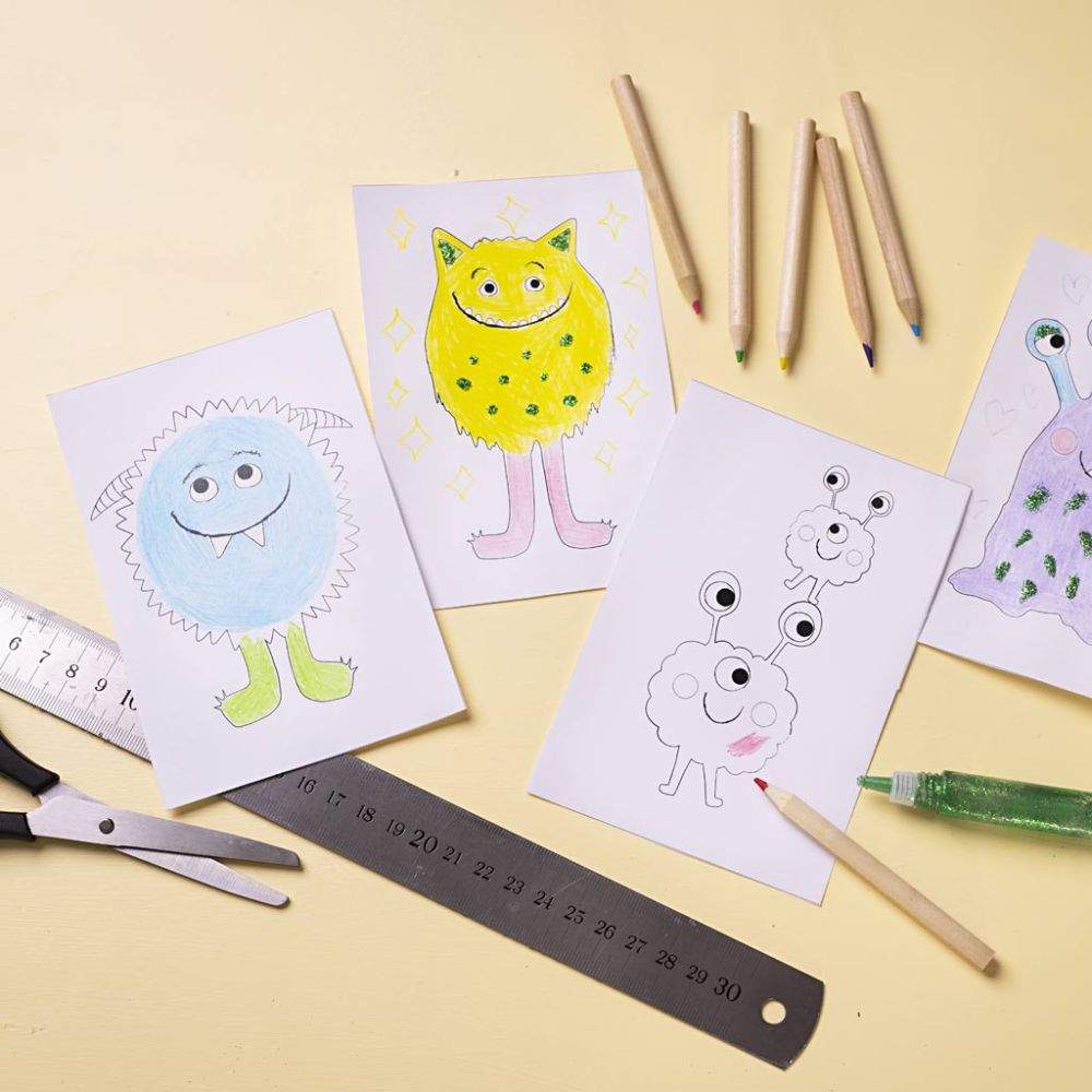 Children's drawings of colorful monsters on paper with art supplies on a yellow background