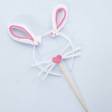 A bunny made from a hairband,  a popsicle stick and pink and white pipe cleaners on a  white background