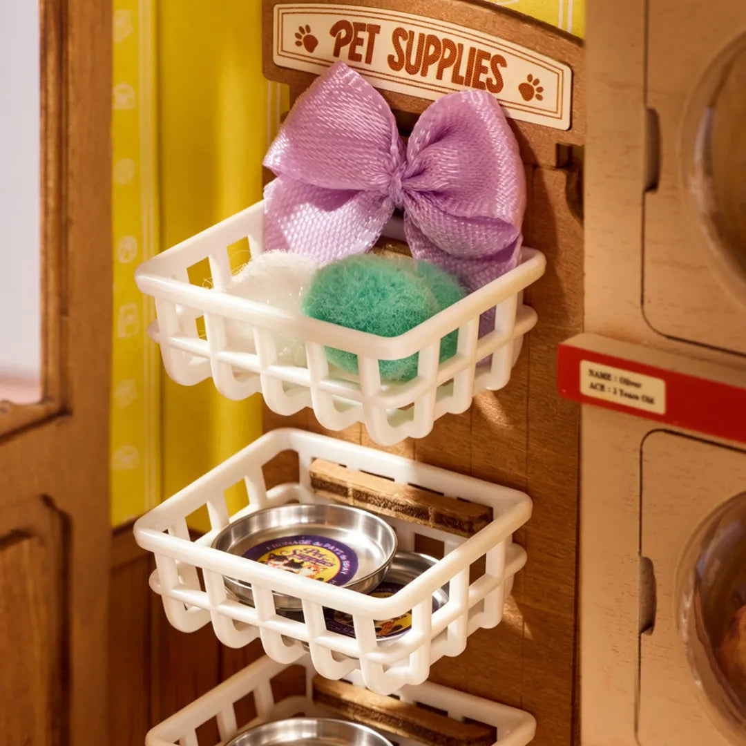 A close-up of a wooden toy play kitchen or pet accessory storage unit with two white plastic basket shelves. The top shelf holds a large lavender bow, white and green cleaning sponges, and a sign reading 'PET SUPPLIES.